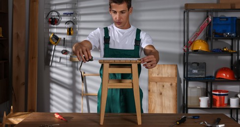 Photo of Man using tape measure while repairing wooden stool indoors