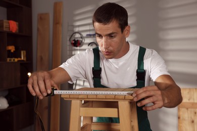 Photo of Man using tape measure while repairing wooden stool indoors