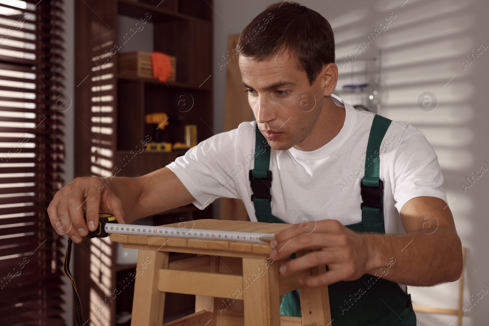 Photo of Man using tape measure while repairing wooden stool indoors