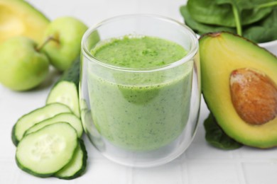 Photo of Tasty green smoothie in glass and products on white tiled table, closeup