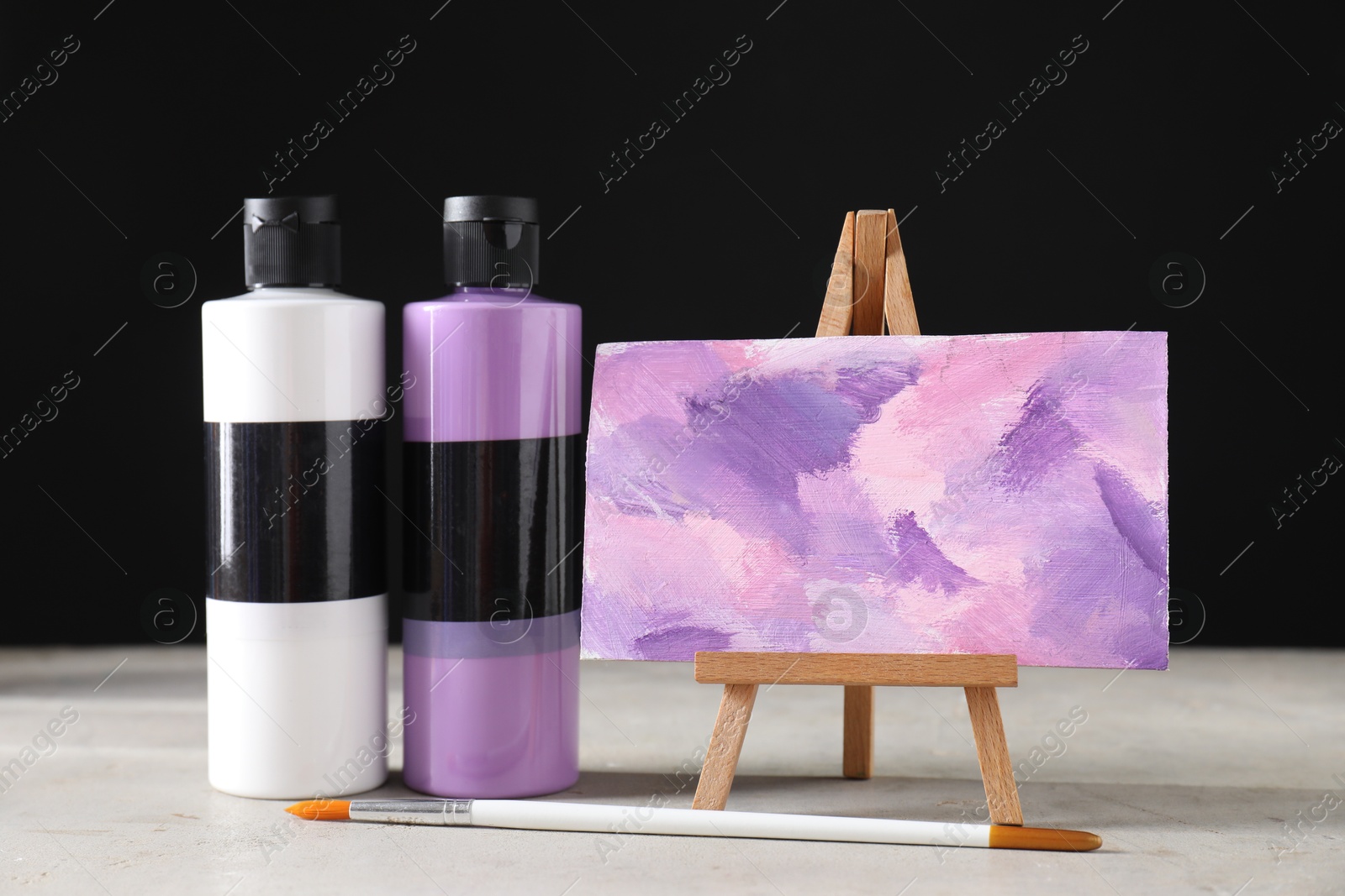 Photo of Acrylic paints of different colors in bottles, brush and small easel with abstract picture on light table