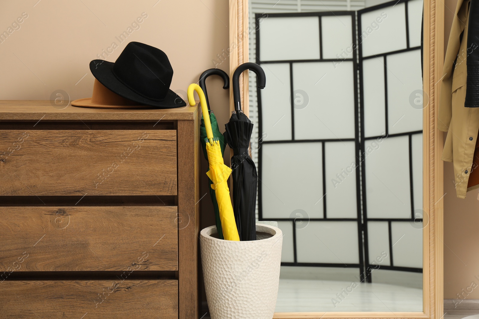 Photo of Colorful umbrellas in holder, hat on wooden chest of drawers and mirror indoors