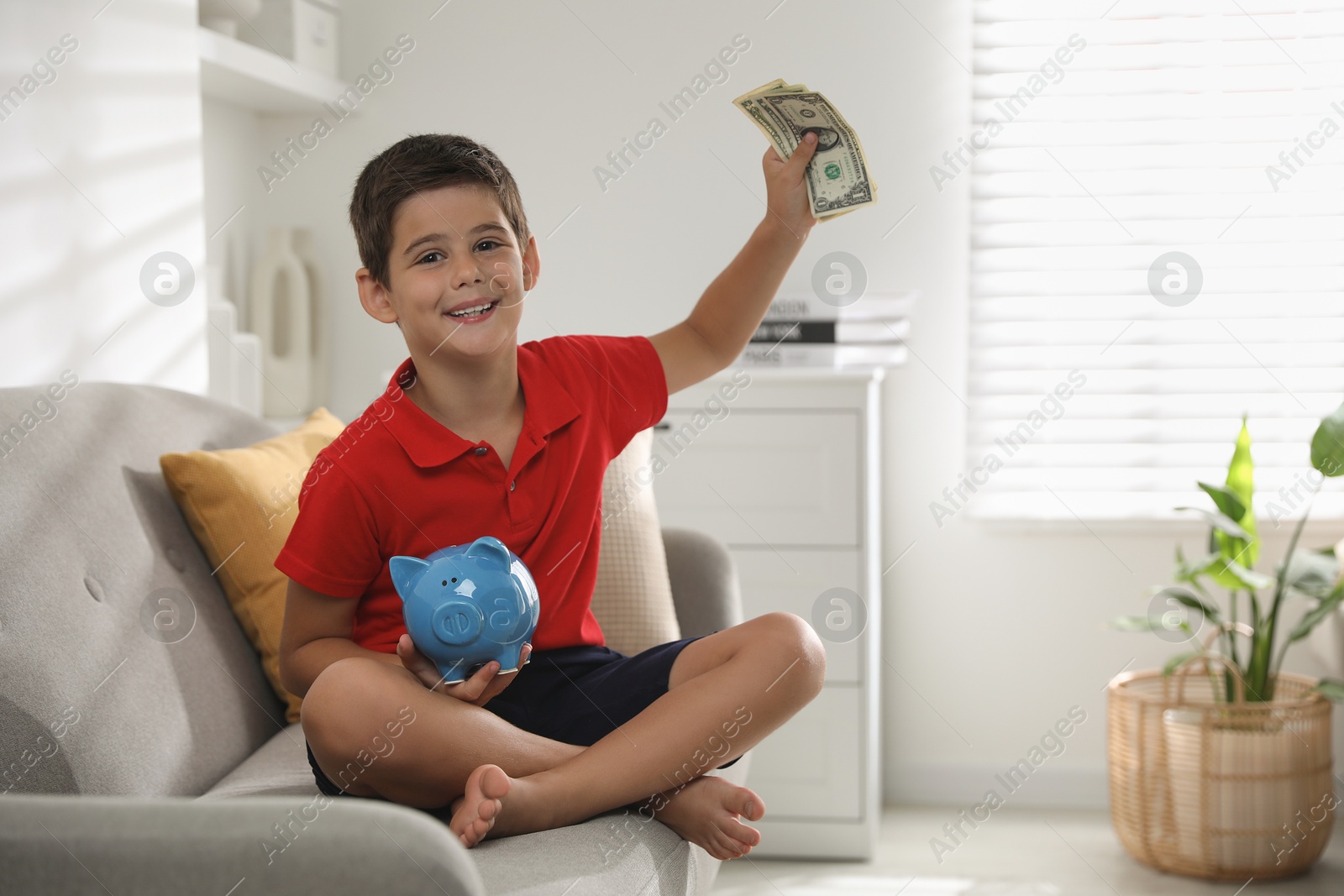 Photo of Cute boy with piggy bank and pocket money on sofa at home, space for text