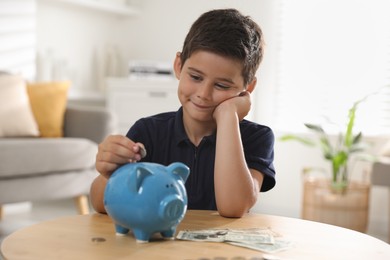 Pocket money. Cute boy putting coin into piggy bank at wooden table indoors