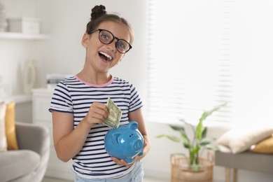 Cute girl with pocket money and piggy bank at home, space for text