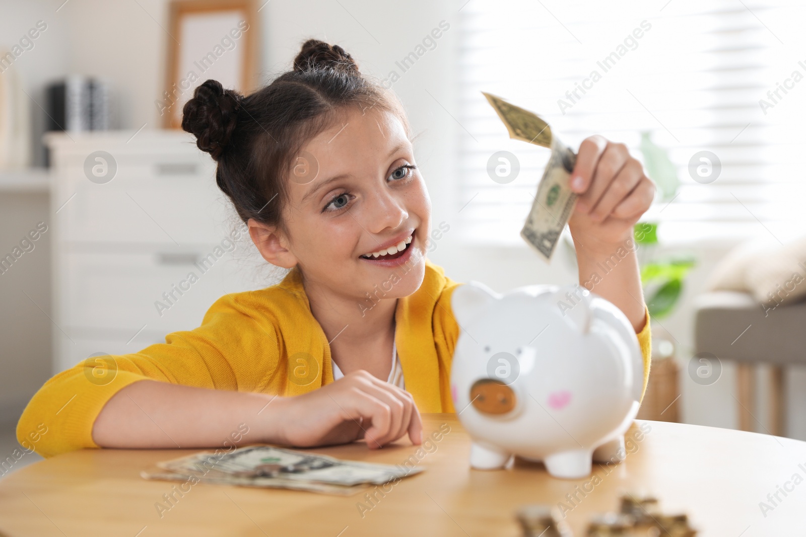 Photo of Pocket money. Cute girl putting dollar banknote into piggy bank at wooden table indoors