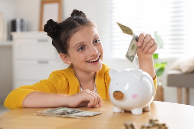 Pocket money. Cute girl putting dollar banknote into piggy bank at wooden table indoors