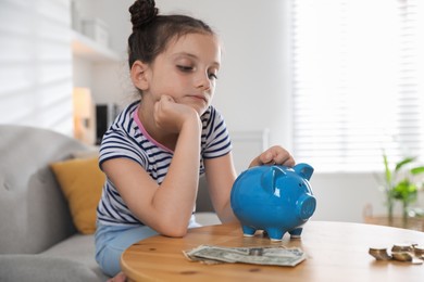 Pocket money. Cute girl putting coin into piggy bank at wooden table indoors