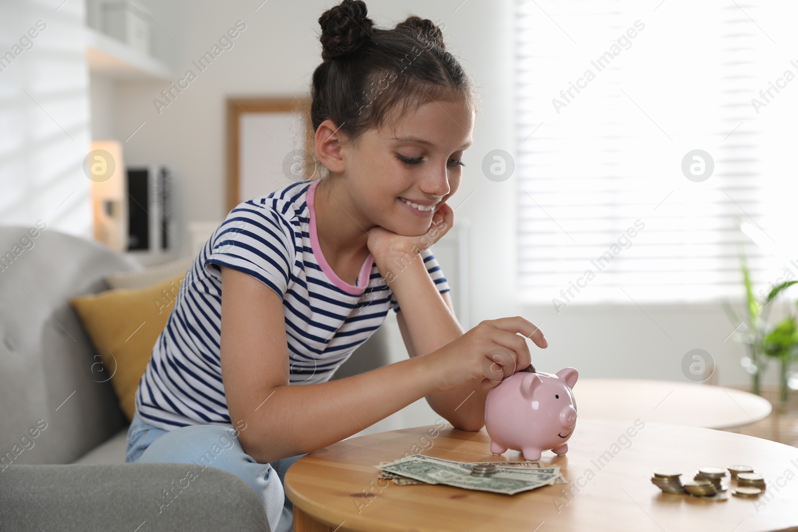 Photo of Pocket money. Cute girl putting coin into piggy bank at wooden table indoors