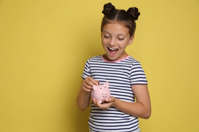 Pocket money. Cute girl putting coin into piggy bank on yellow background