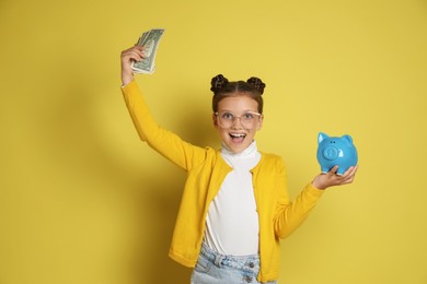 Photo of Pocket money. Cute girl with piggy bank and dollar banknotes on yellow background