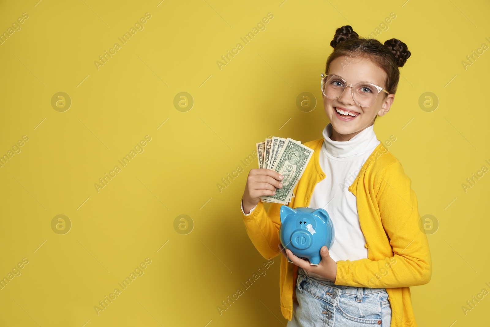 Photo of Pocket money. Cute girl with piggy bank and dollar banknotes on yellow background, space for text