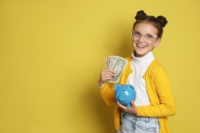 Photo of Pocket money. Cute girl with piggy bank and dollar banknotes on yellow background, space for text