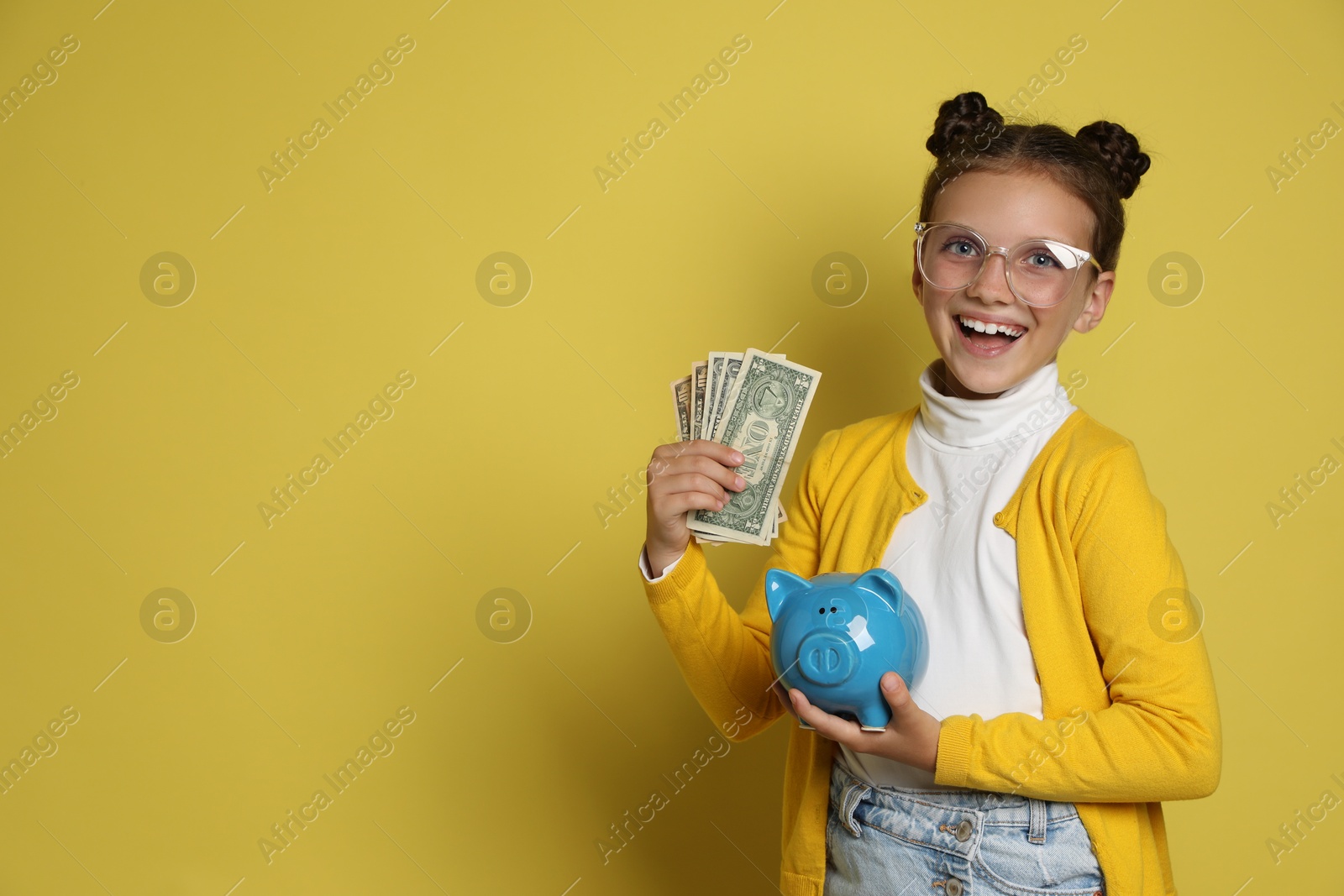 Photo of Pocket money. Cute girl with piggy bank and dollar banknotes on yellow background, space for text