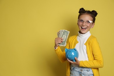 Photo of Pocket money. Cute girl with piggy bank and dollar banknotes on yellow background, space for text