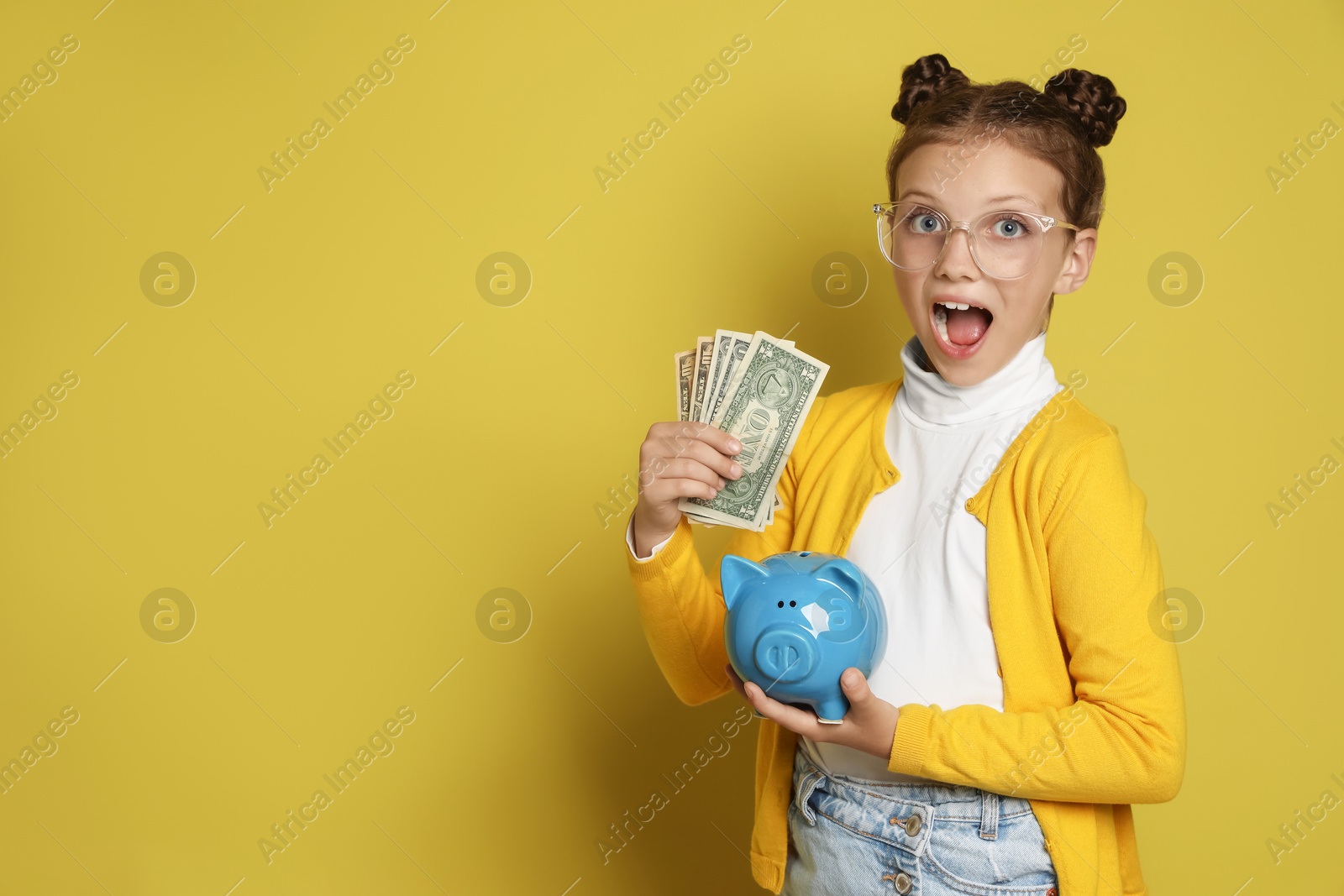 Photo of Pocket money. Cute girl with piggy bank and dollar banknotes on yellow background, space for text