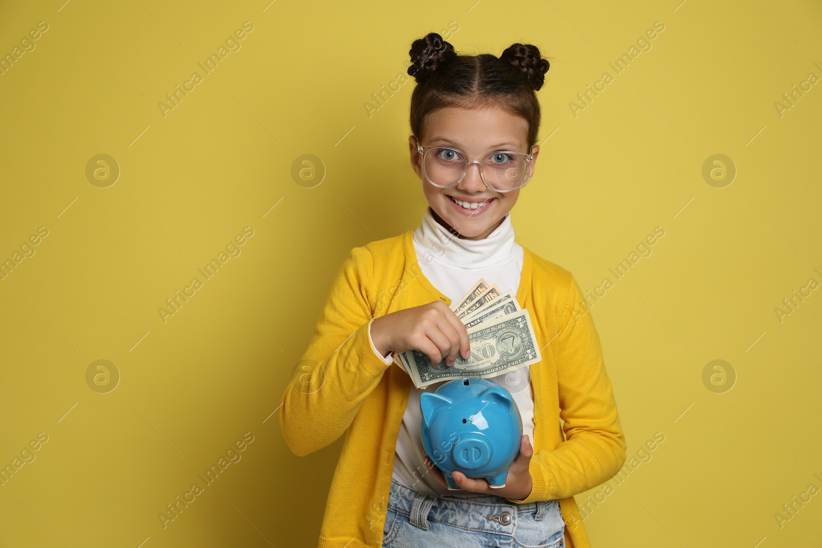 Photo of Pocket money. Cute girl with piggy bank and dollar banknotes on yellow background