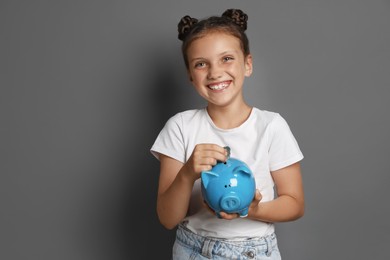 Pocket money. Cute girl putting coins into piggy bank on grey background