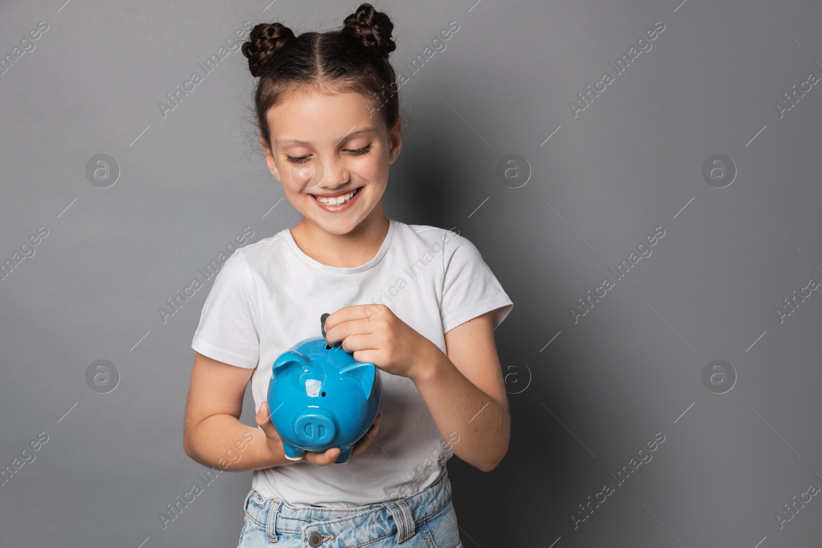 Photo of Pocket money. Cute girl putting coins into piggy bank on grey background