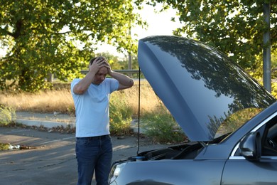 Stressed man looking under hood of broken car outdoors