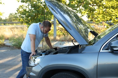 Stressed man looking under hood of broken car outdoors