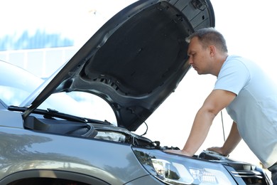 Stressed man looking under hood of broken car outdoors