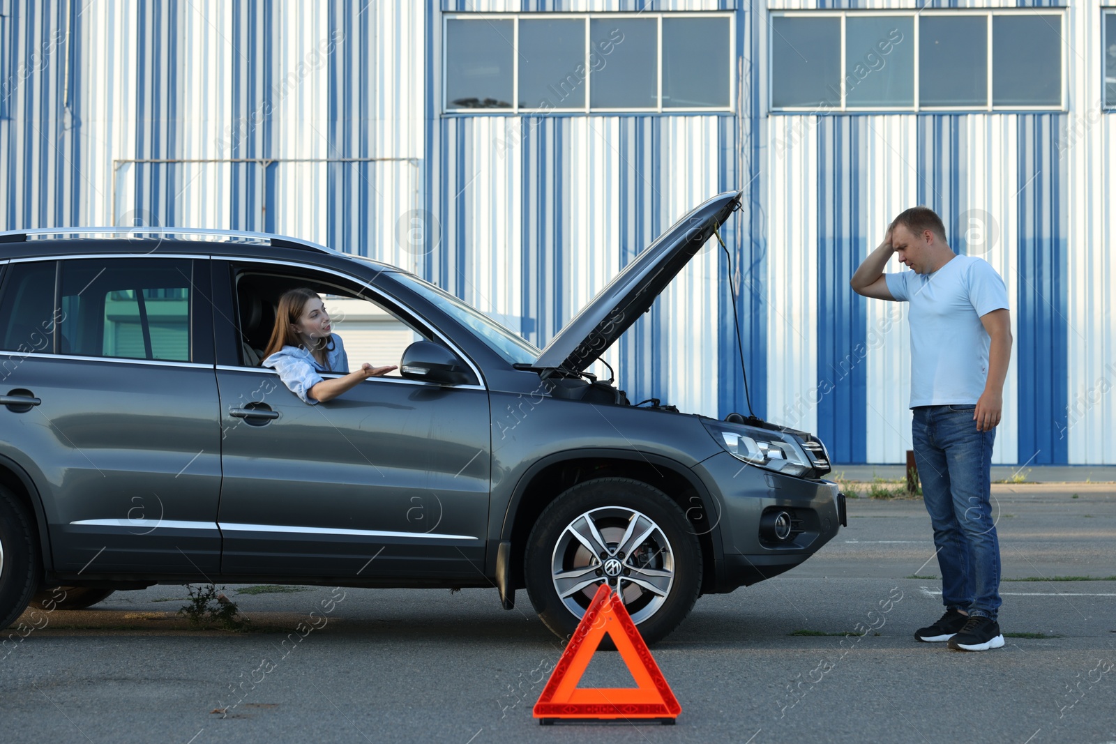 Photo of Stressed man standing near broken car while woman sitting in auto outdoors