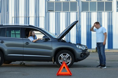 Stressed man standing near broken car while woman sitting in auto outdoors