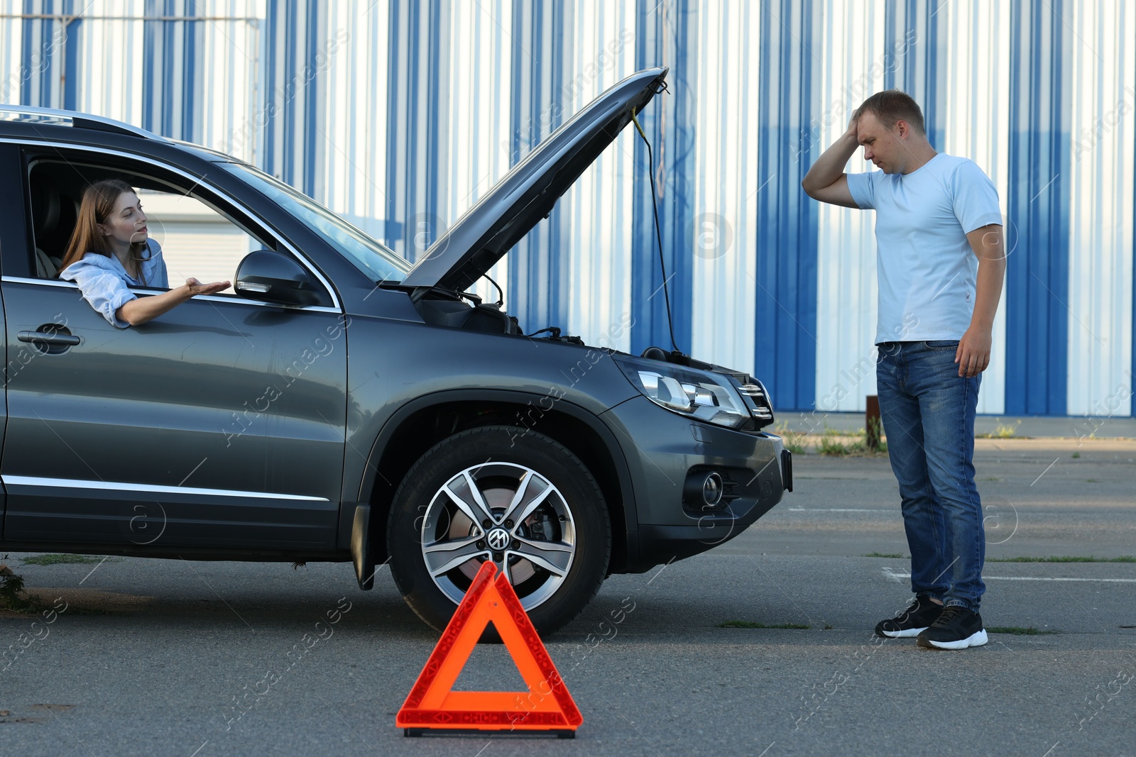 Photo of Stressed man standing near broken car while woman sitting in auto outdoors