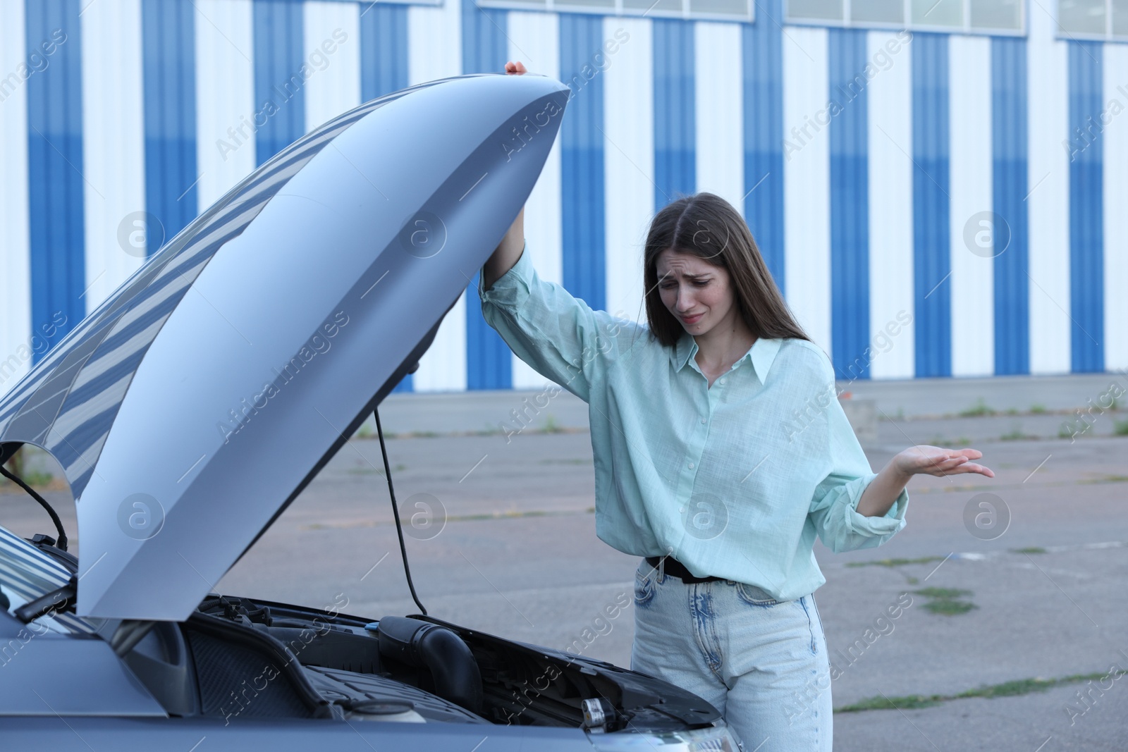 Photo of Stressed woman looking under hood of broken car outdoors