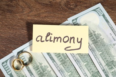 Card with word Alimony, dollar banknotes and rings on wooden table, closeup