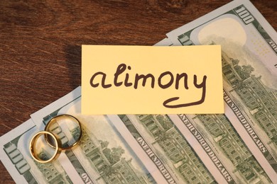 Card with word Alimony, dollar banknotes and rings on wooden table, closeup