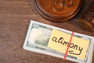 Photo of Card with word Alimony, dollar banknotes, gavel and rings on wooden table, above view