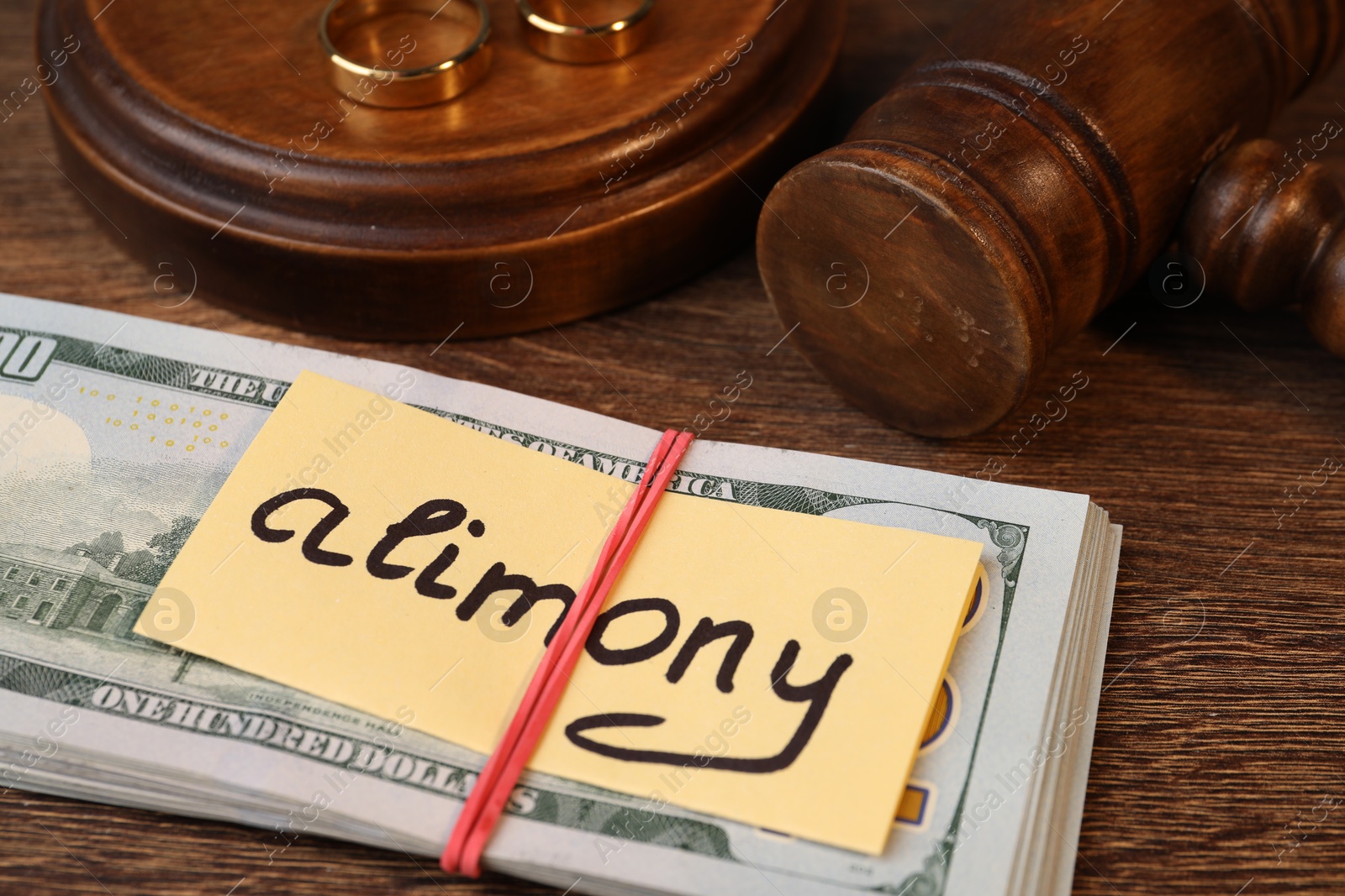 Photo of Card with word Alimony, dollar banknotes, gavel and rings on wooden table, closeup