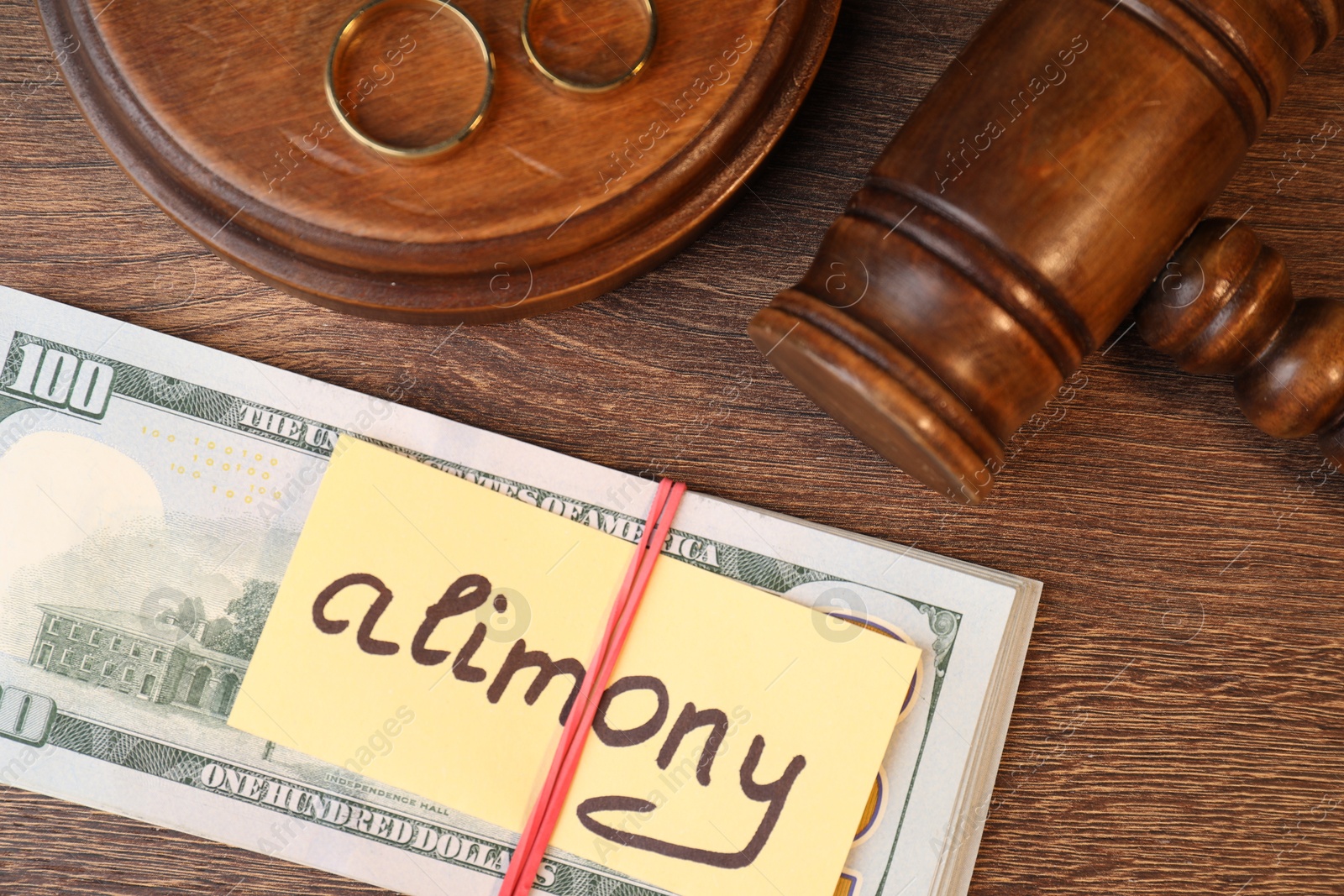 Photo of Card with word Alimony, dollar banknotes, gavel and rings on wooden table