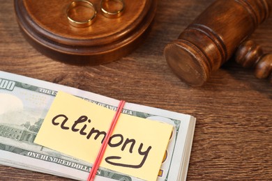 Photo of Card with word Alimony, dollar banknotes, gavel and rings on wooden table, closeup