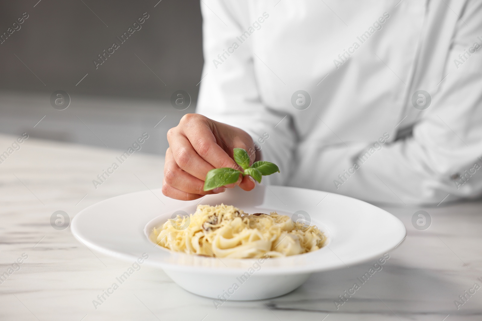 Photo of Professional chef decorating delicious pasta with basil at white marble table in kitchen