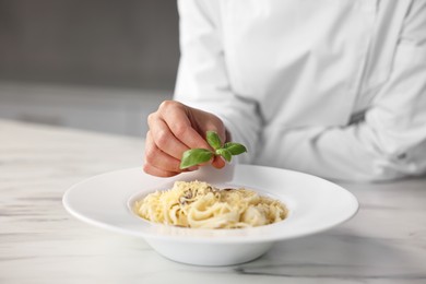 Photo of Professional chef decorating delicious pasta with basil at white marble table in kitchen