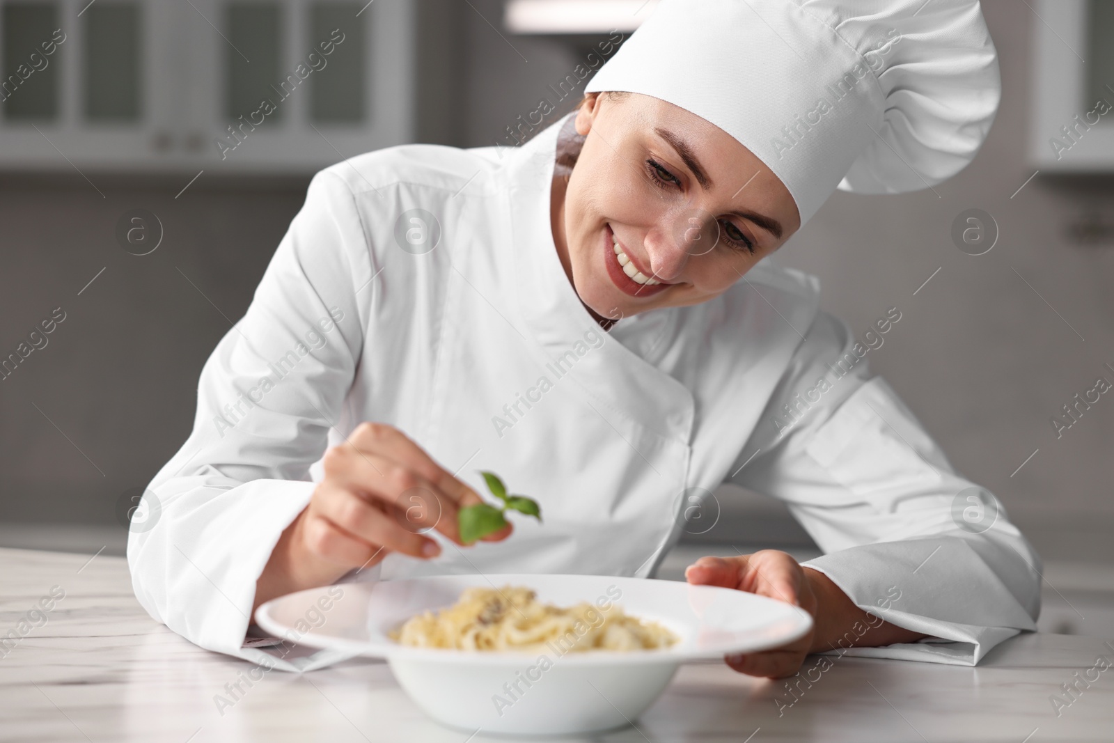 Photo of Professional chef decorating delicious pasta with basil at white marble table in kitchen