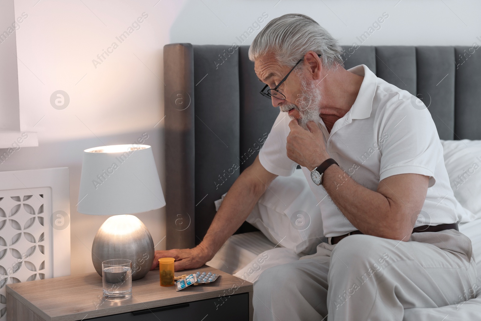 Photo of Senior man near bedside table with pills and glass of water indoors