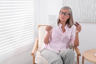 Senior woman with pills reading medicine instruction in armchair at home, space for text