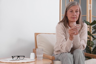 Photo of Senior woman holding blister with pills in armchair at home, space for text