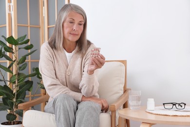 Senior woman holding blister with pills in armchair at home, space for text