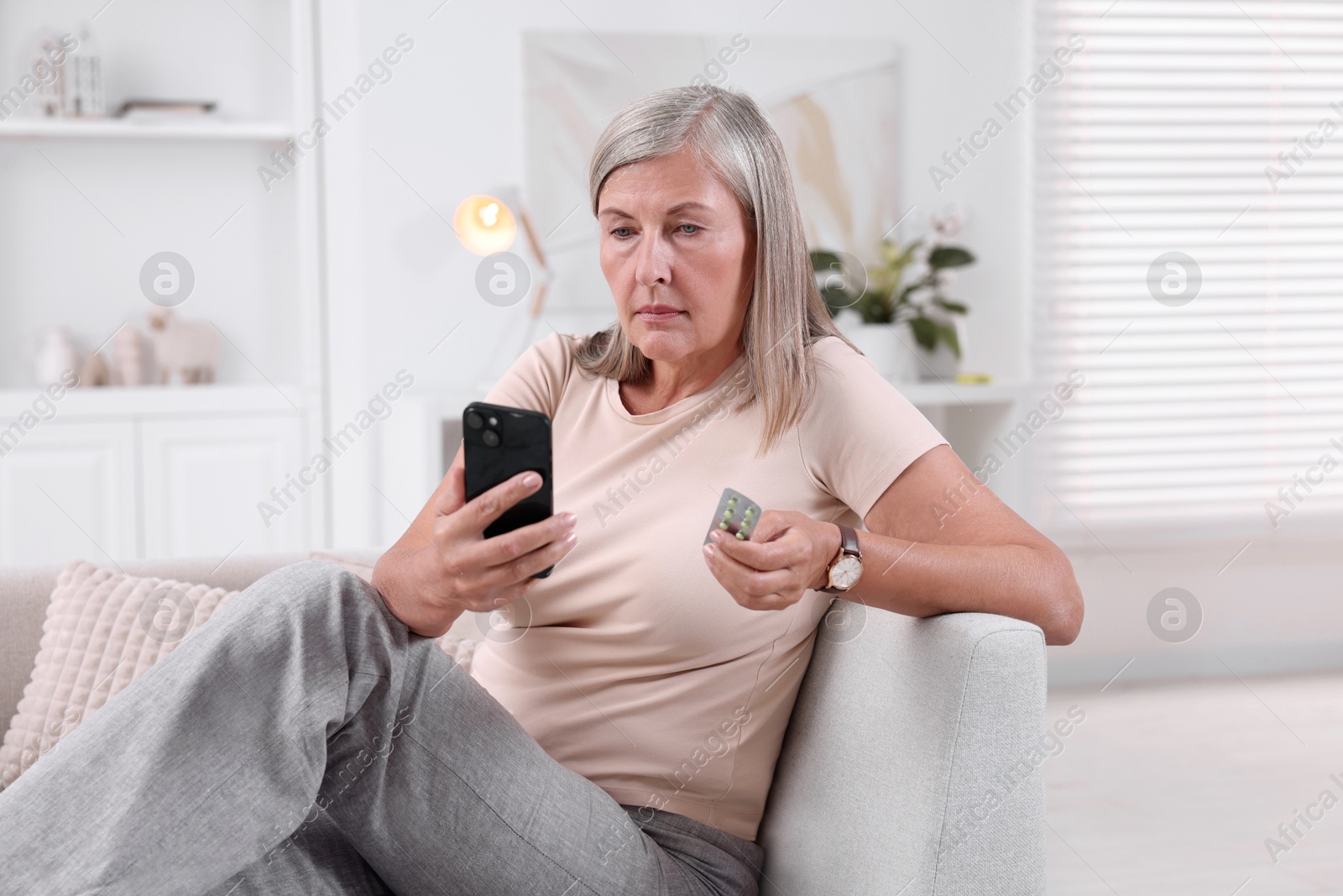 Photo of Senior woman with pills using smartphone on sofa at home