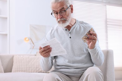 Senior man with pills reading medicine instruction on sofa at home