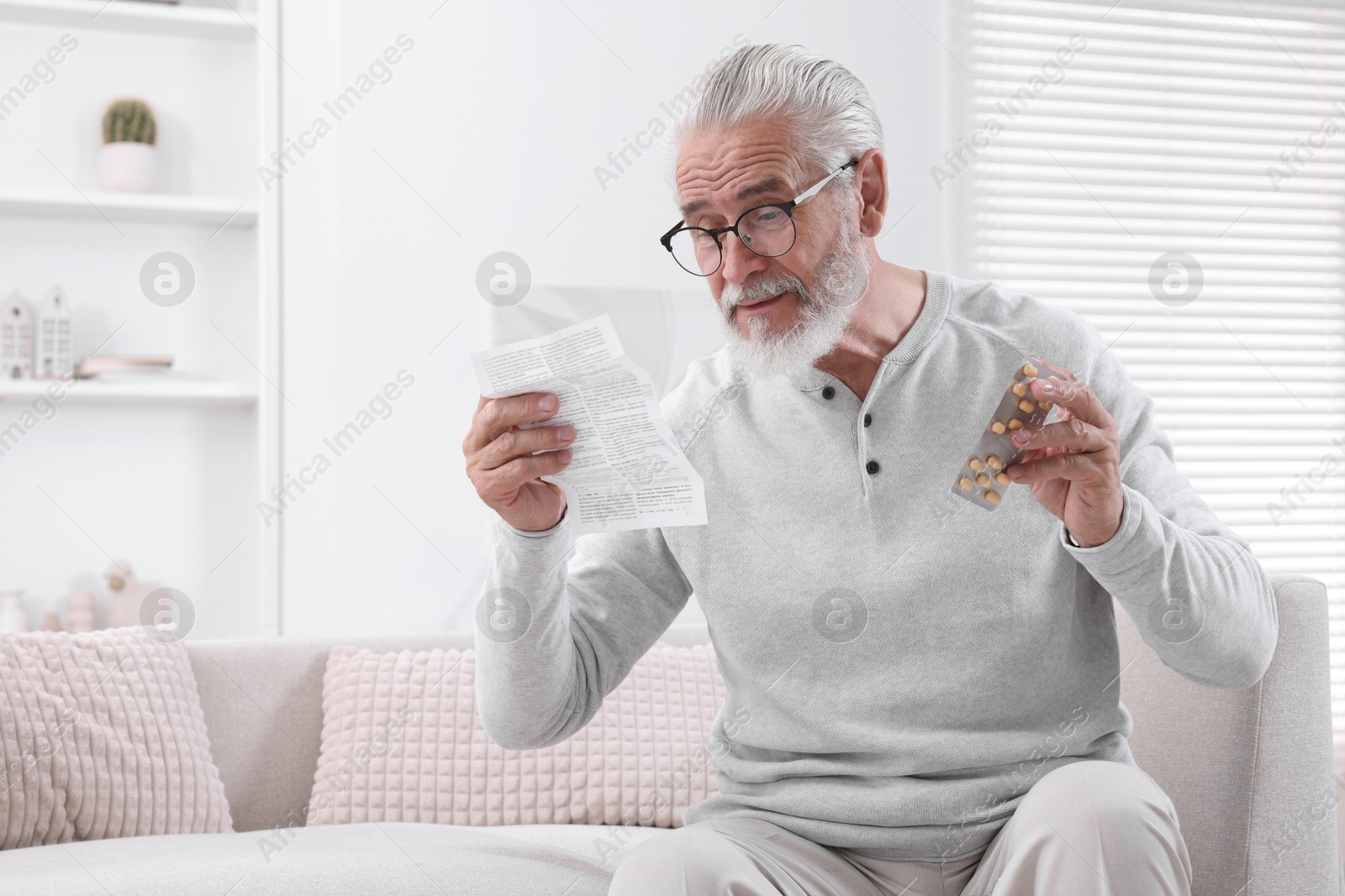 Photo of Senior man with pills reading medicine instruction on sofa at home