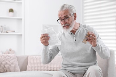 Senior man with pills reading medicine instruction on sofa at home