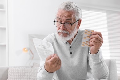 Photo of Senior man with pills reading medicine instruction on sofa at home