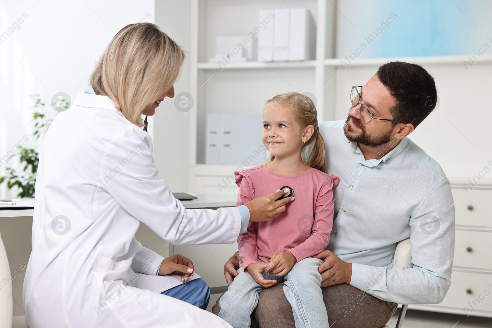 Photo of Doctor examining little girl with stethoscope and her father in hospital