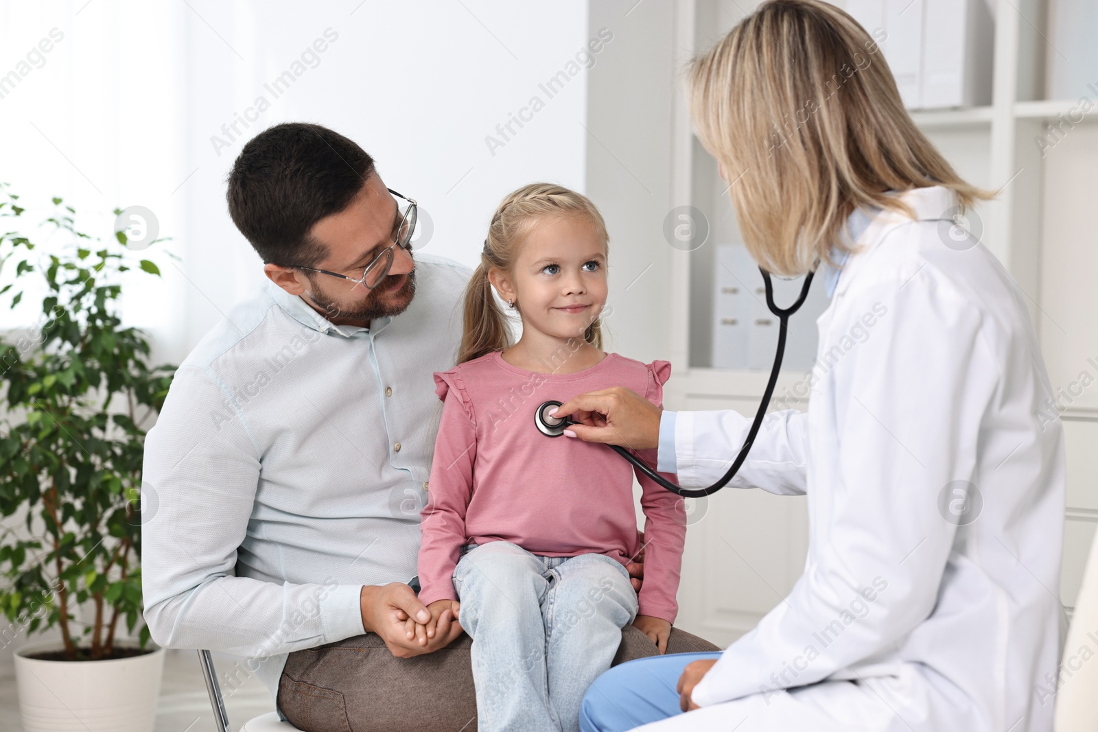 Photo of Doctor examining little girl with stethoscope and her father in hospital